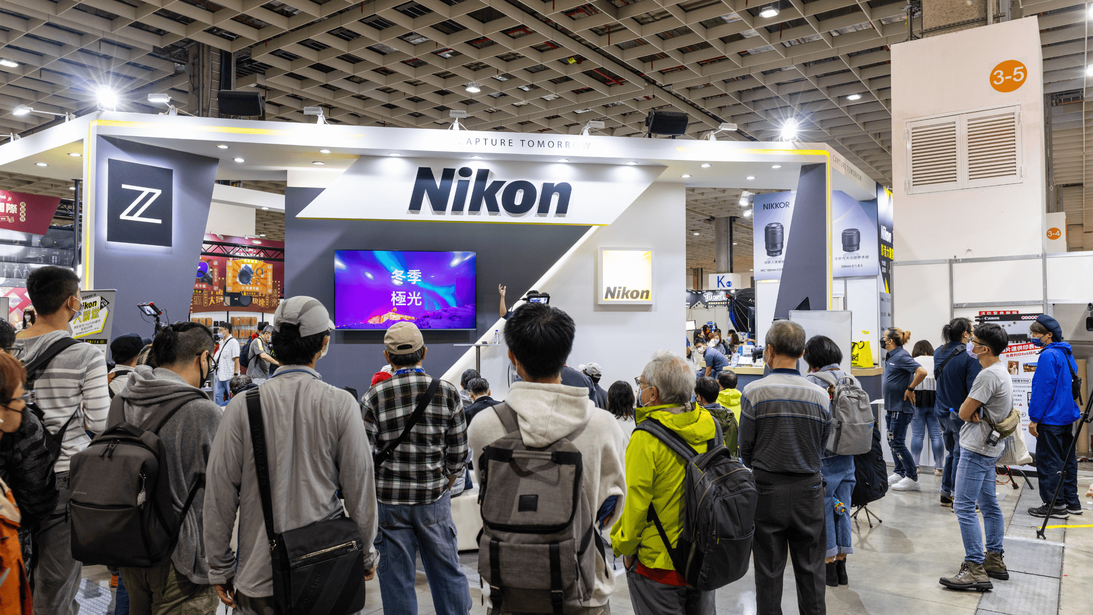 A crowd of attendees gathered around a trade show booth, watching a live presentation in an exhibit hall.