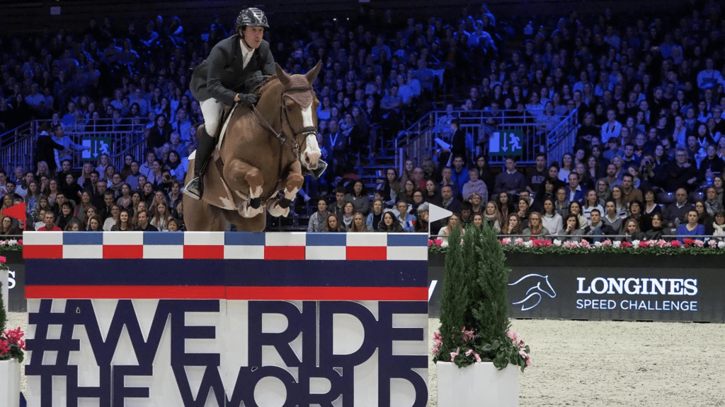 A horse and rider clearing a fence at the Longines Masters, with the stadium crowd behind.