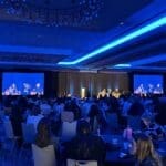 Conference attendees seated at round tables in a hotel ballroom, facing a stage with two large screens showing a panel discussion.